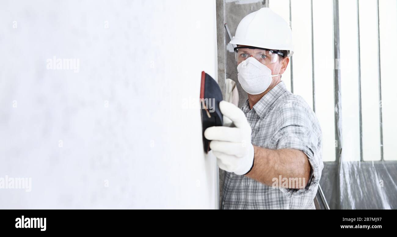 homme pondu le mur avec du papier de verre, travailleur de la construction professionnelle avec masque, casque de sécurité, gants et lunettes de protection. site de bâtiment intérieur, Banque D'Images
