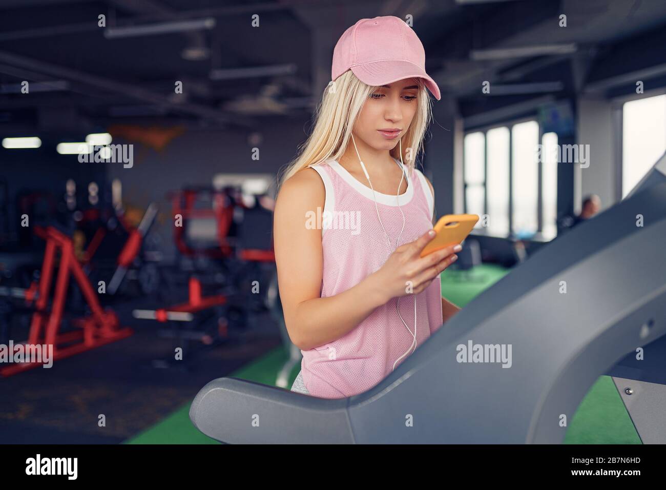 Vue latérale d'une femme blonde sportive dans une casquette rose se  préparant à faire de l'exercice sur tapis roulant en choisissant de la  musique sur smartphone dans la salle de sport Photo