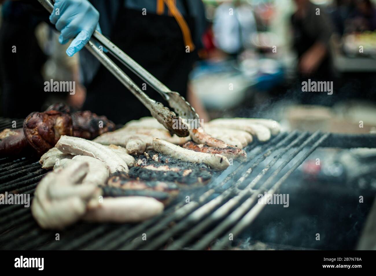 Vue rapprochée avec peu de profondeur de champ sur les mains d'un chef à l'aide de pinces pour préparer et griller des saucisses dans un étalage de nourriture de rue pendant le marché agricole. Banque D'Images