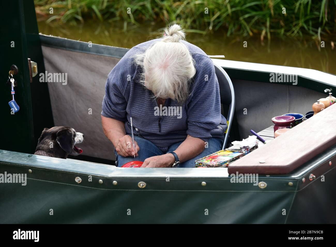 Femme aux cheveux gris peignant un article, regardé par son chien, sur la poupe d'un narrowboat pendant un festival de canal à Whitchurch, Shropshire Royaume-Uni Banque D'Images