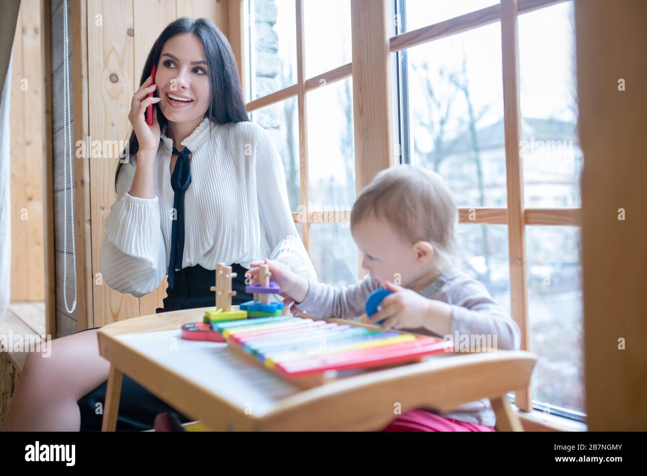 Jeune maman assise sur windowsill, parler au téléphone, sourire, jouer avec la fille Banque D'Images