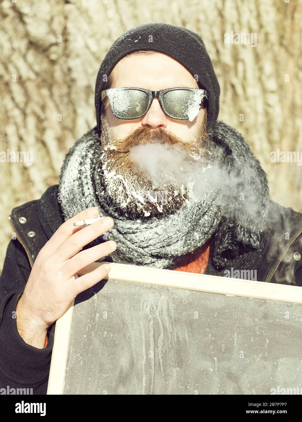 Beau homme, hipster, avec barbe et moustache dans des lunettes de soleil  noires couvertes de givre blanc fume cigarette tenant blanc tableau noir ou  tableau de surveillance le jour d'hiver sur fond