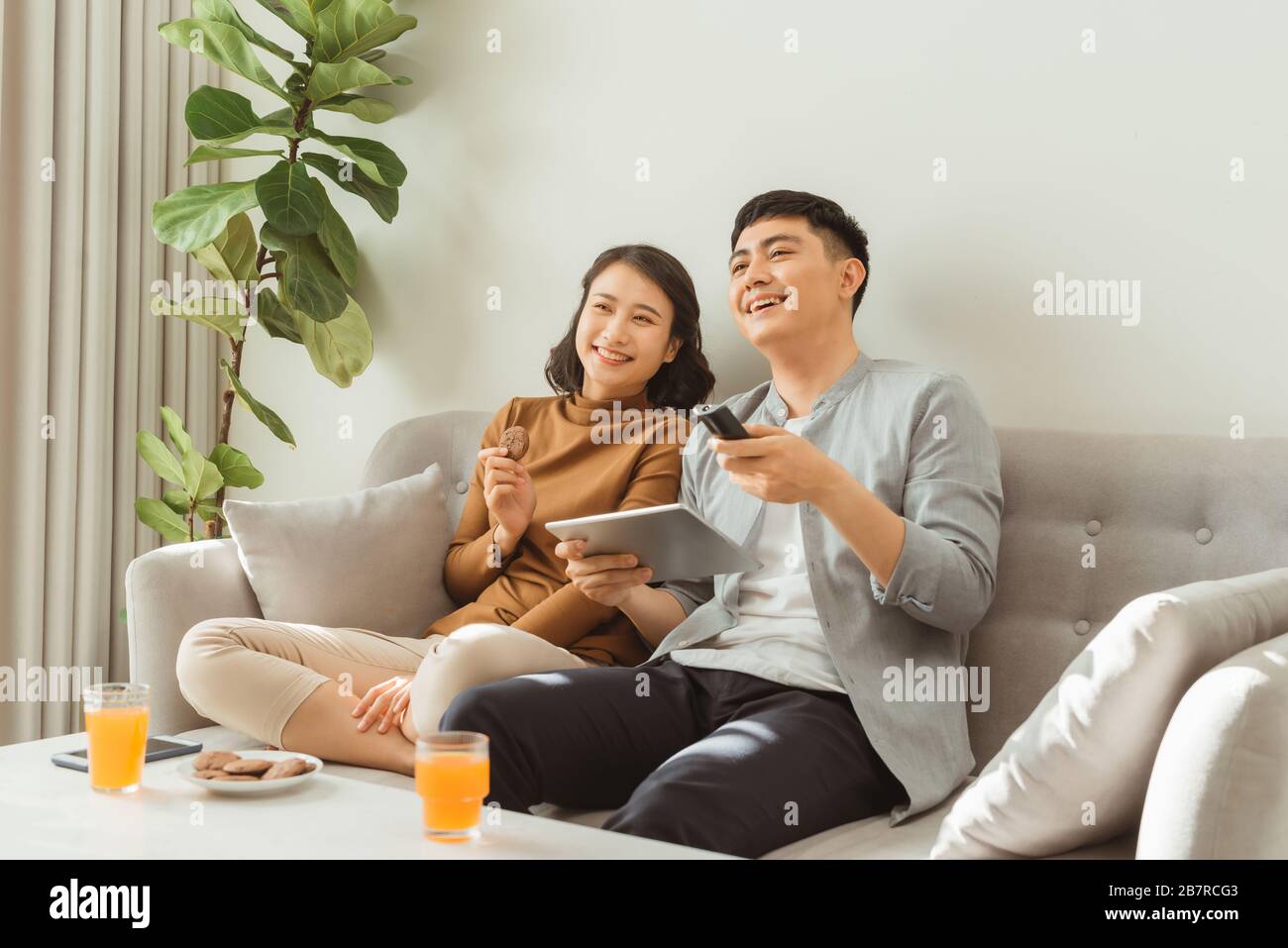 Couple watching tv assis sur un canapé dans le salon à la maison. L'homme est le zapping et woman is holding a tablet and looking at you Banque D'Images