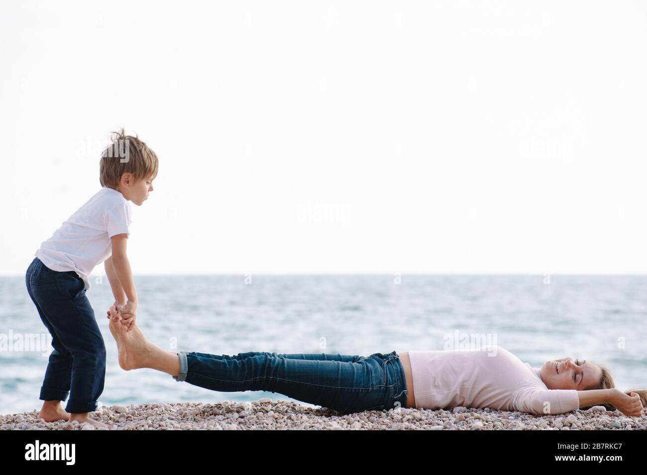 drôle actif petit garçon soulevant sa mère allongé sur les jambes de plage de mer pendant le printemps loisirs de loisirs en plein air jeux Banque D'Images