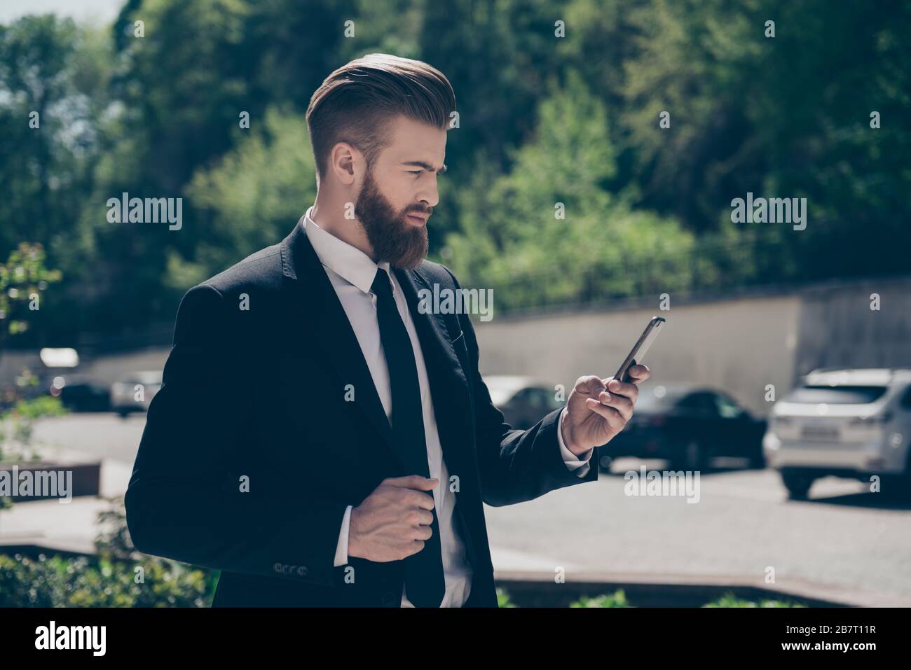 Jeune homme d'affaires barbu élégant dans un costume classe parcourt son pda dans la rue le jour ensoleillé. Il commande une cabine par pda Banque D'Images