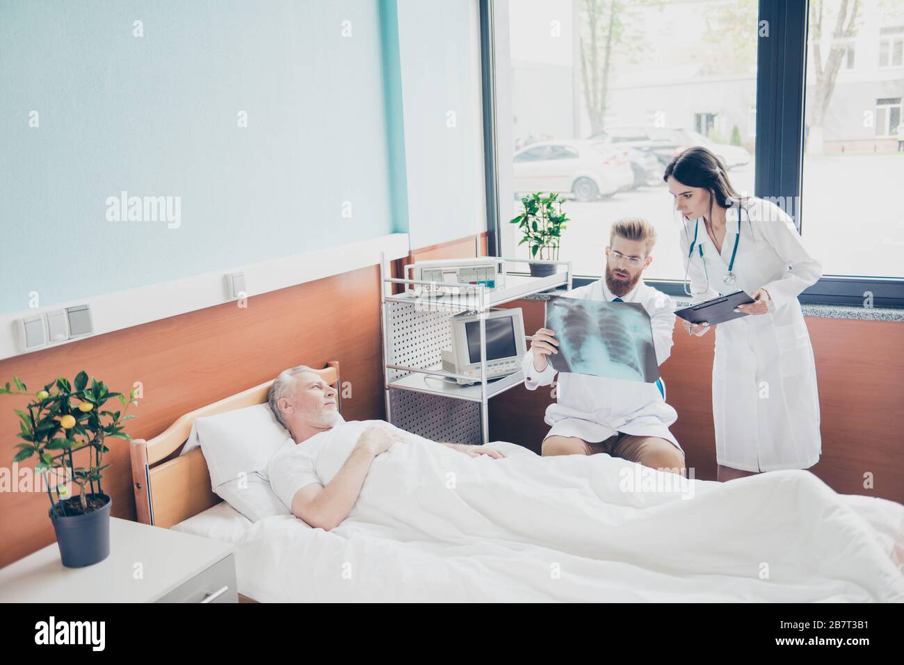 Le jeune doc barbu rouge montre la radiographie à l'infirmière et parle de l'opération, le patient est âgé, il est dans le lit bouleversé Banque D'Images