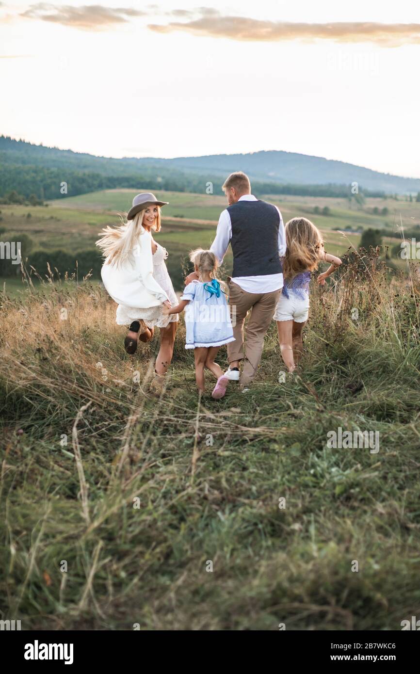 Enchantée, la famille Cauasian se réjouit de courir dans le champ vert d'été au coucher du soleil. Jolie jeune femme mère qui tourne en arrière et regarde l'appareil photo. Marchez Banque D'Images