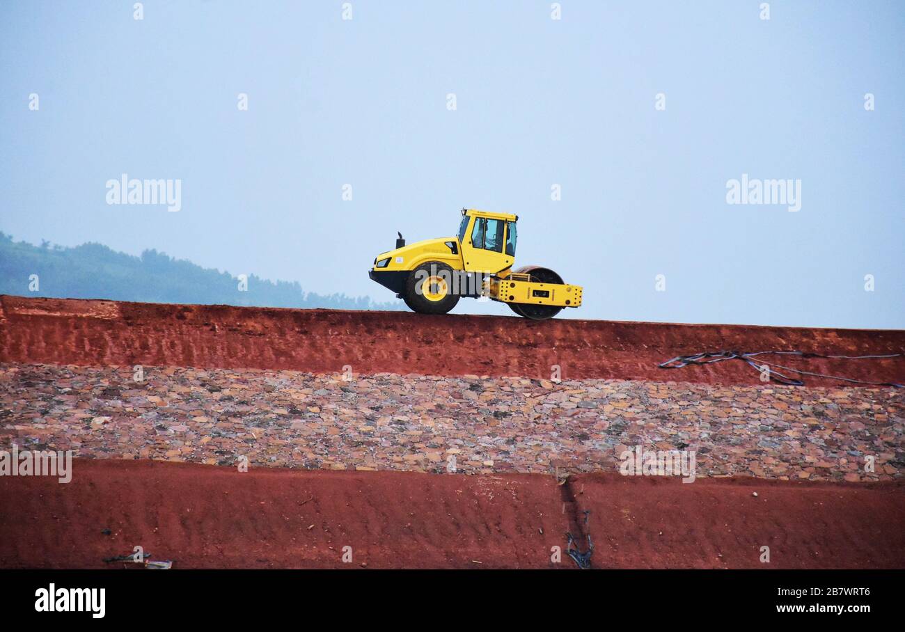 Machine à rouleaux de route jaune compactant le sol sur un site de construction de barrage avec ciel bleu Banque D'Images