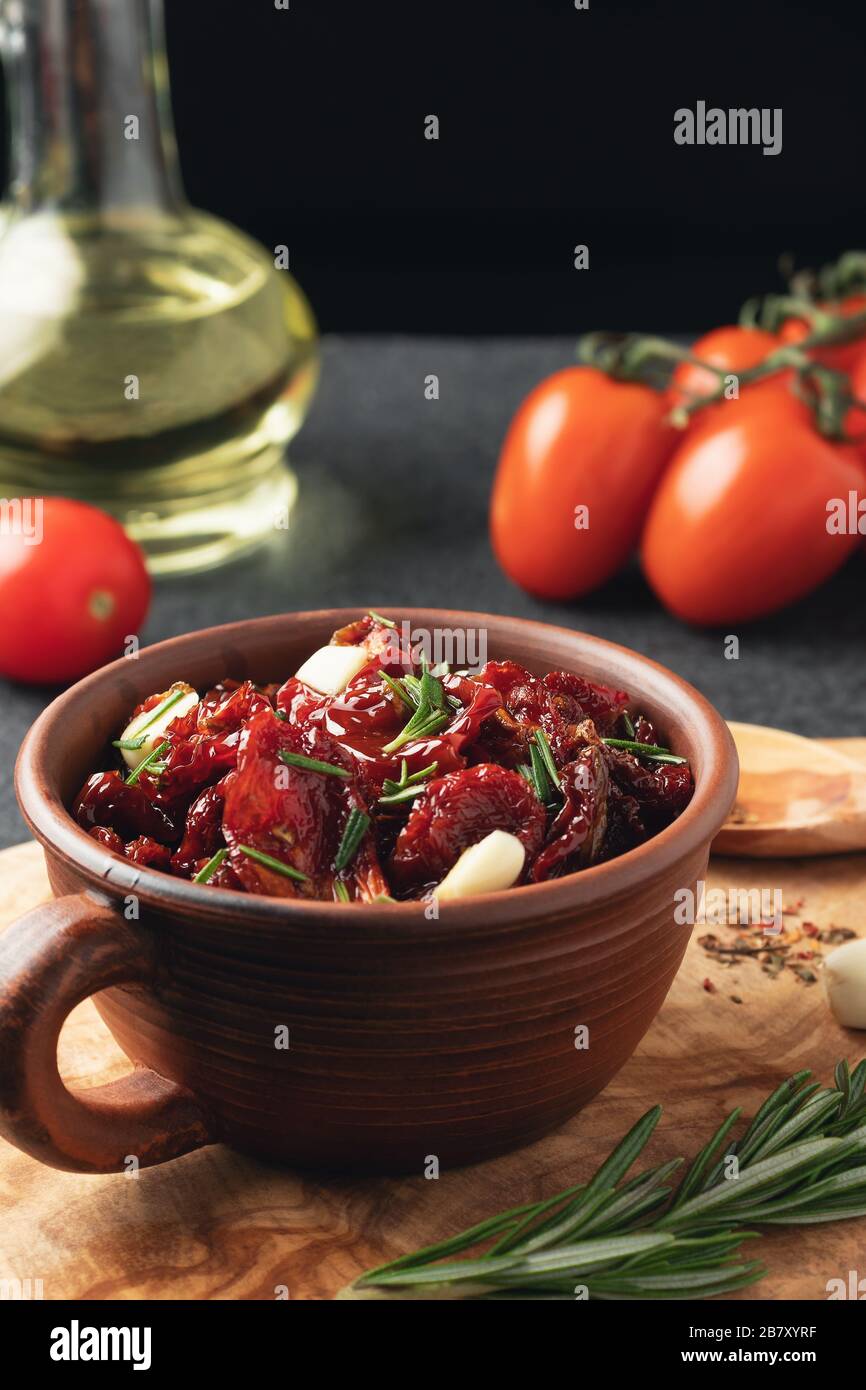 Tomates séchées au soleil avec ail, romarin et épices dans un bol en argile sur un tableau de découpe en bois d'olive, image verticale. Banque D'Images