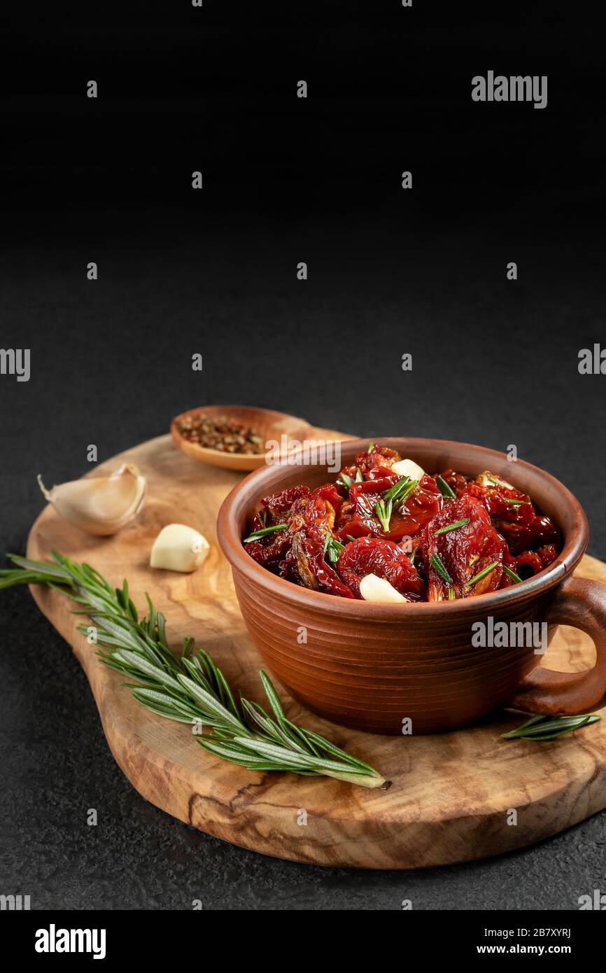 Tomates séchées au soleil avec ail, romarin et épices dans un bol en argile sur un tableau de découpe en bois d'olive, image verticale avec espace copyspace. Banque D'Images