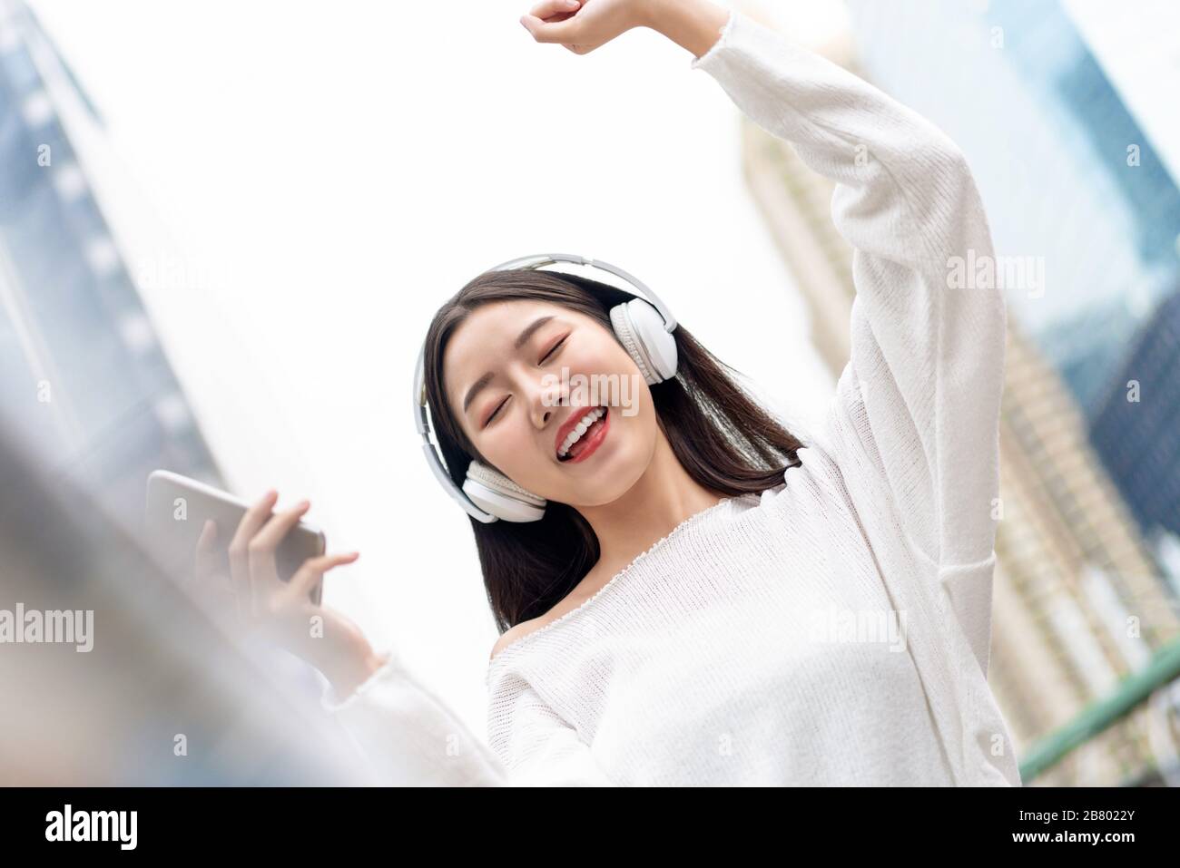 Bonne fille asiatique souriante portant un casque et écoutant de la musique en streaming avec des yeux fermés sur le fond du bâtiment de la ville Banque D'Images
