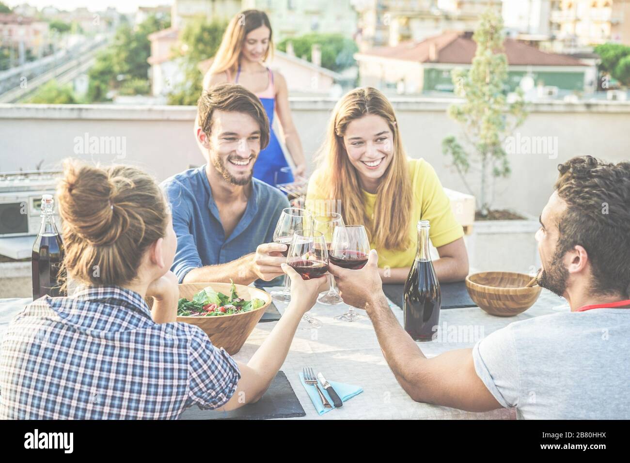 Des amis joyeux applaudissent avec du vin rouge à la fête barbecue sur le toit - des gens heureux qui font un dîner barbecue en plein air avec vue sur la ville en arrière-plan - Focus on bott Banque D'Images