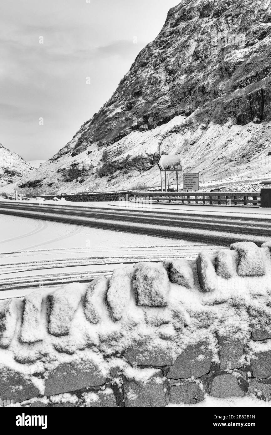 Conduisez sur la gauche dans différentes langues et les panneaux des attractions touristiques sur la route dans Glencoe couvert de neige, Highlands, Ecosse en hiver - noir et blanc Banque D'Images