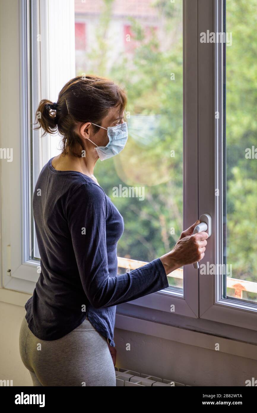 Une femme mise en quarantaine à la maison, portant un masque médical et une tenue décontractée, est en veille devant une fenêtre fermée, en regardant dans l'espace. Banque D'Images