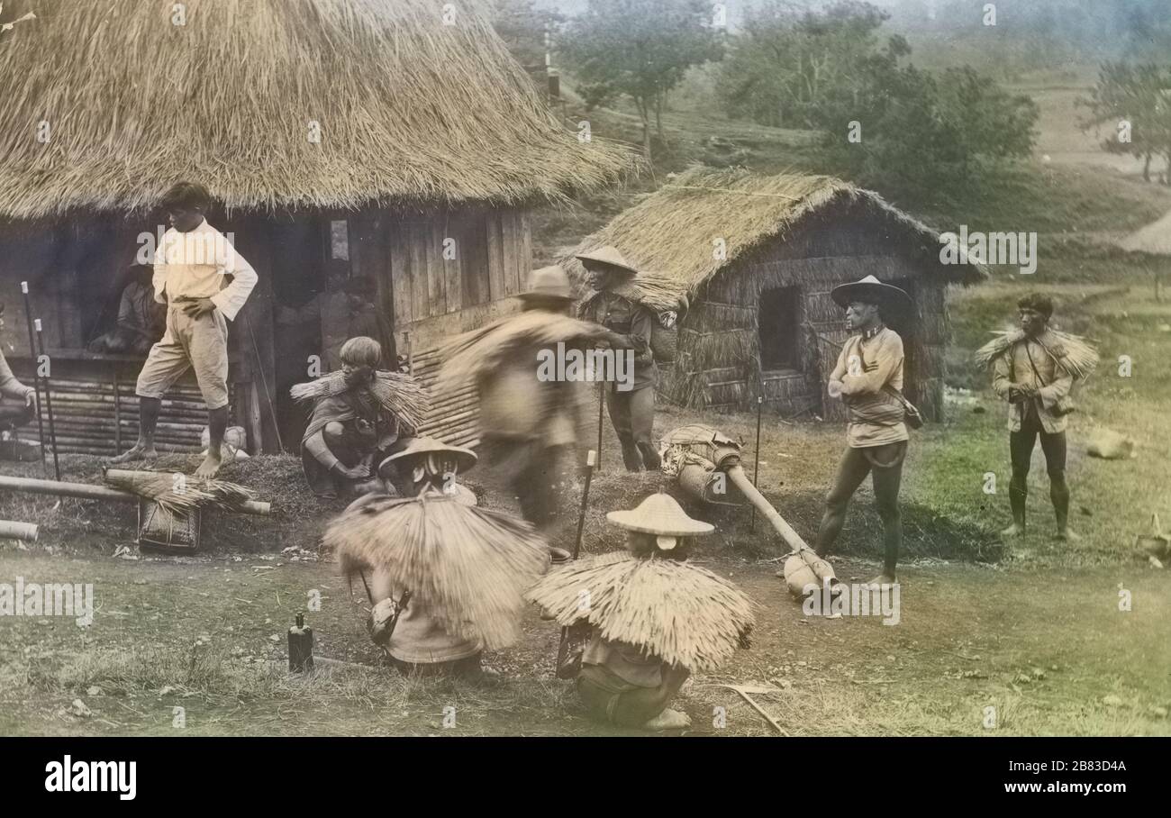 Carte postale monochrome d'un groupe d'hommes, certains portant des capes de pluie en chaume, devant les huttes de nipa, l'île de Luzon, les Philippines, 1915. De la bibliothèque publique de New York. Remarque : l'image a été colorisée numériquement à l'aide d'un processus moderne. Les couleurs peuvent ne pas être précises sur une période donnée. () Banque D'Images