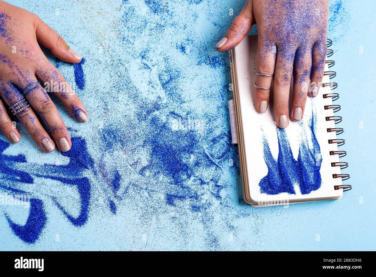 Feuille blanche vide d'ordinateur portable avec la main de la femme au-dessus du stylo, et poudre bleue décorative autour sur un fond bleu pastel, espace de copie. Banque D'Images