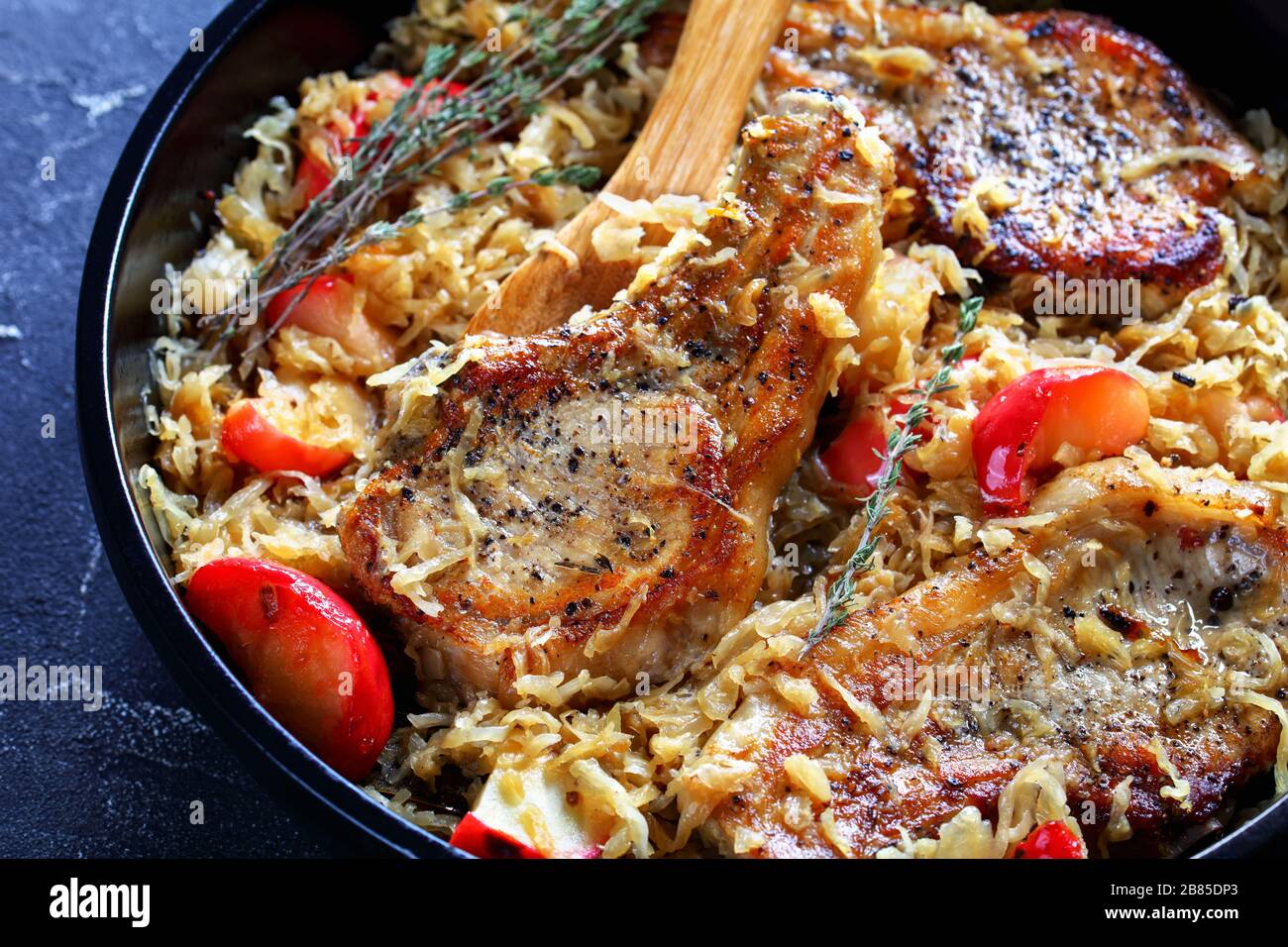 Gros plan de côtelettes de porc avec Sauerkraut et pommes sur une cuillère en bois dans une braiser en céramique noire sur une table en béton, cuisine allemande, vue horizontale fr Banque D'Images