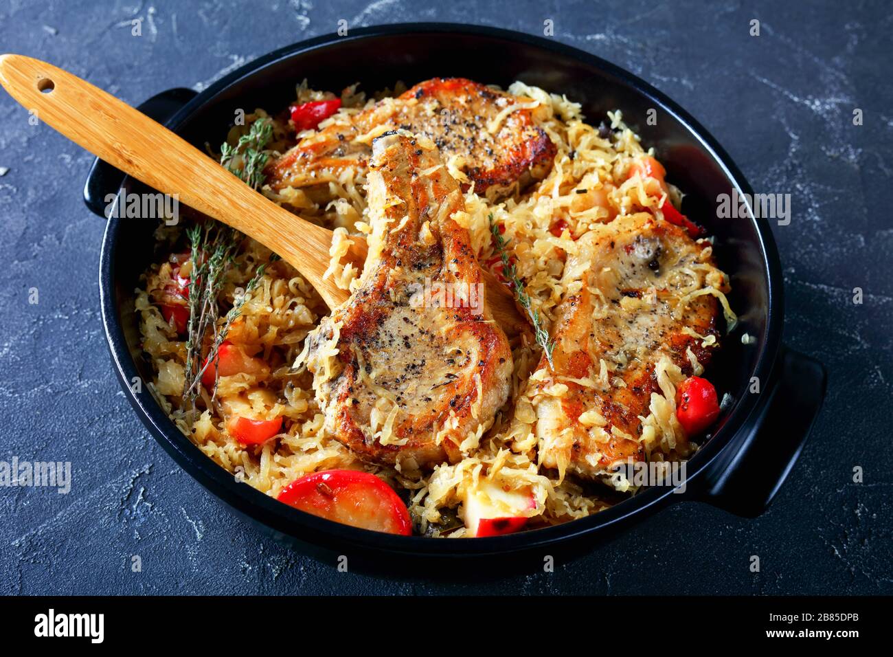 Gros plan de côtelettes de porc avec Sauerkraut et pommes sur une cuillère en bois dans une braiser en céramique noire sur une table en béton, cuisine allemande, vue horizontale f Banque D'Images