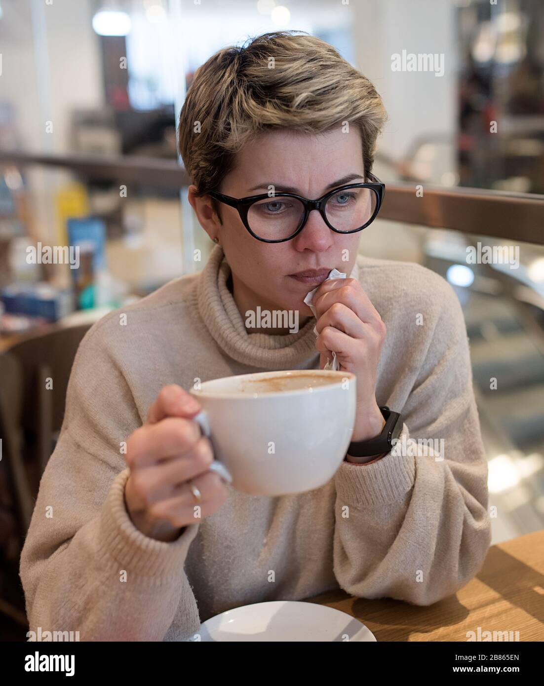 Femme portant un pull ivoire et des verres noirs, téléphone à pointes, essuyant son nez et avoir une tasse de thé après avoir fait du shopping dans un café Banque D'Images