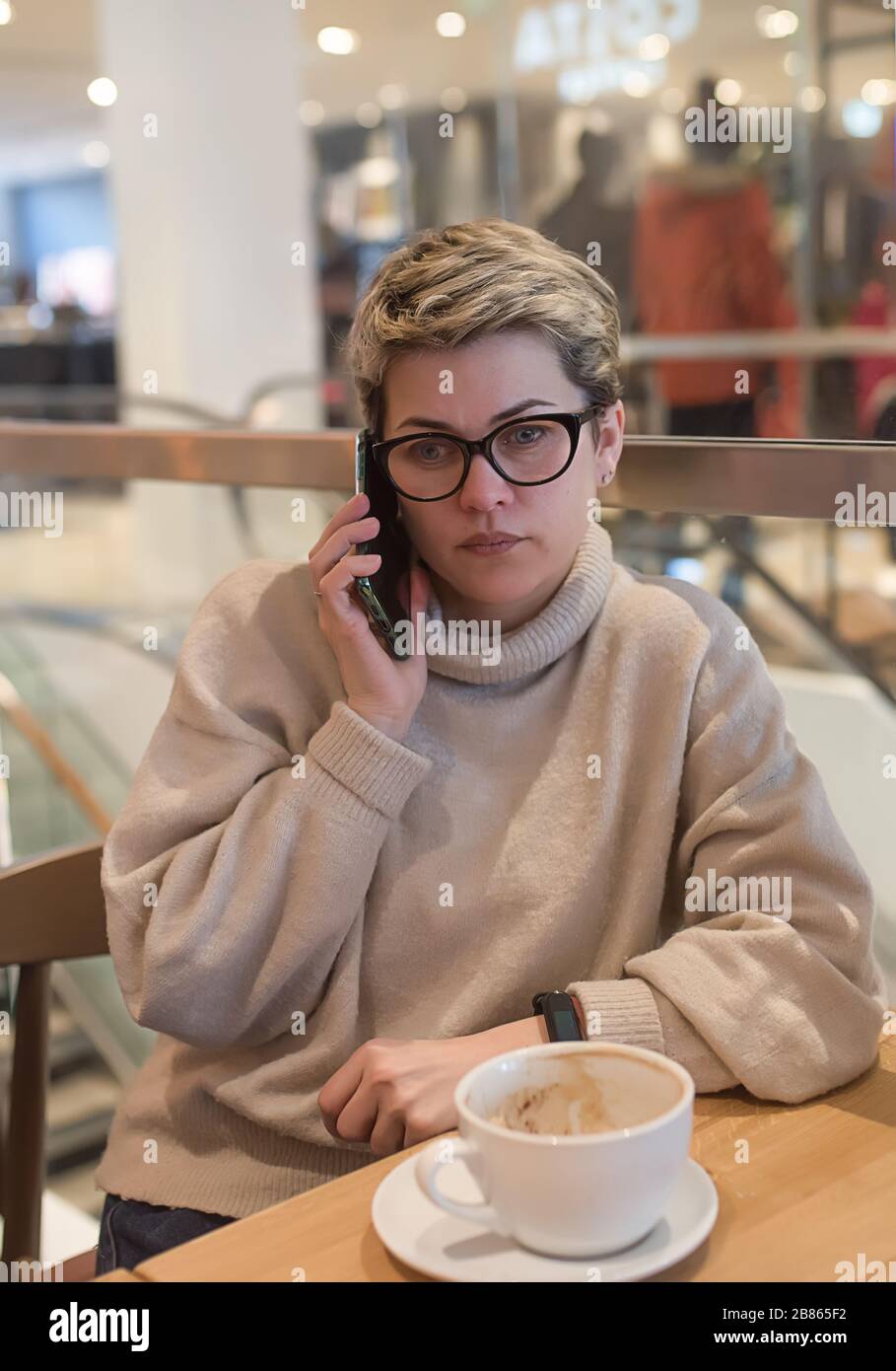 Femme portant un pull ivoire et des verres noirs, téléphone à pointes, essuyant son nez et avoir une tasse de thé après avoir fait du shopping dans un café Banque D'Images