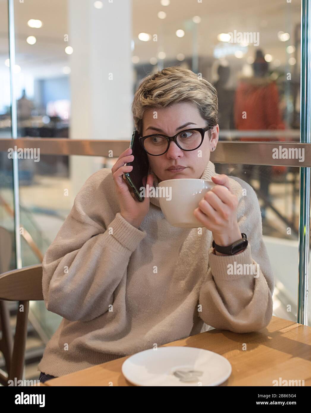 Femme portant un pull ivoire et des verres noirs, téléphone à pointes, essuyant son nez et avoir une tasse de thé après avoir fait du shopping dans un café Banque D'Images