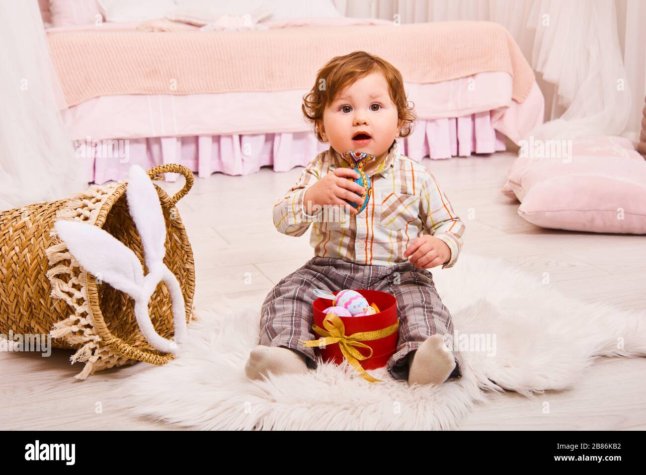 Rire bébé garçon avec des oreilles de lapin chassant pour des œufs de Pâques à la maison. Mignon petit garçon à poil dur assis sur le sol avec une boîte d'œufs de Pâques Banque D'Images