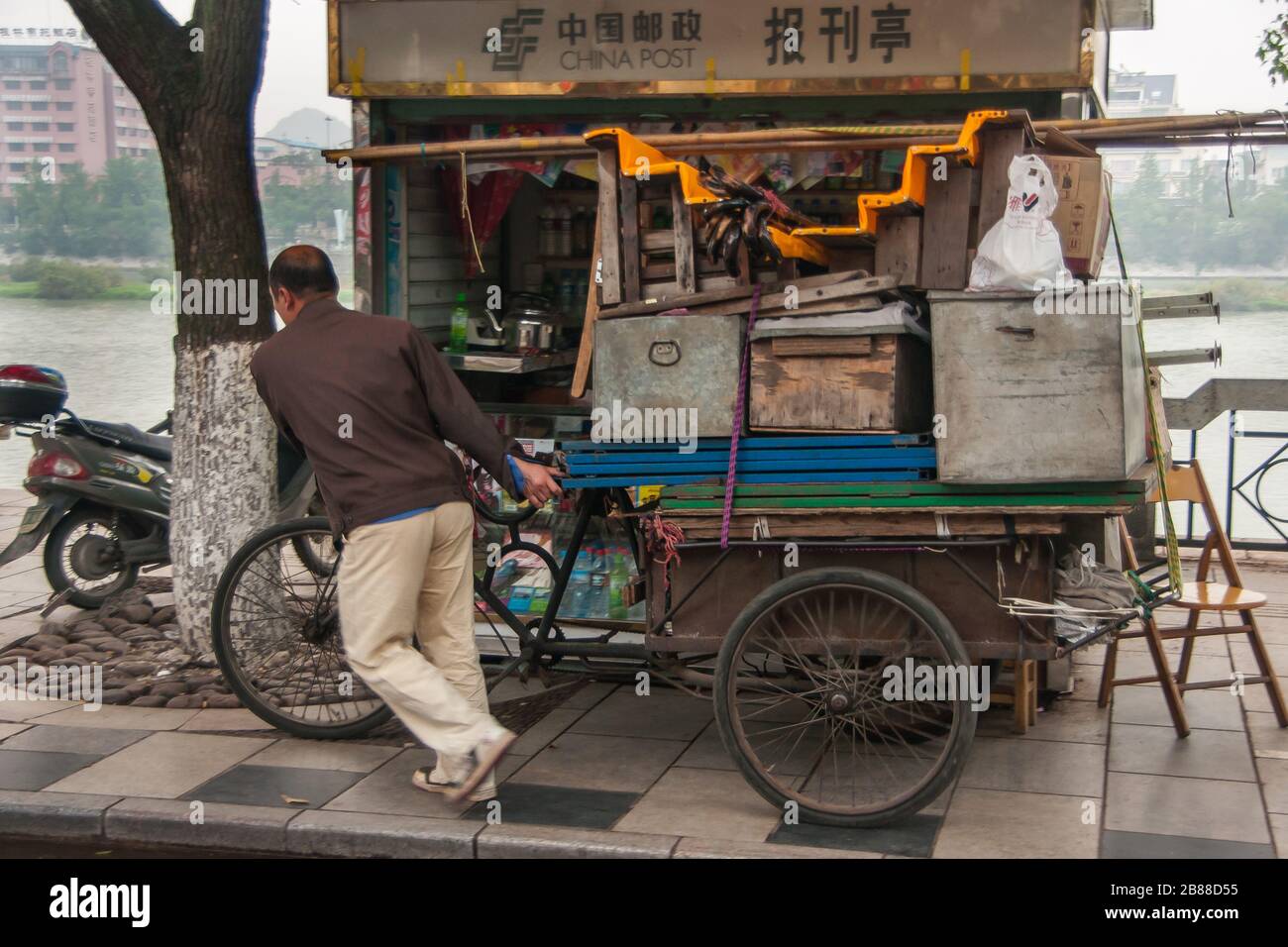 Guilin, Chine - 11 mai 2010: Centre-ville. Gros plan de l'homme tirant surchargée vieux vélo de tricycle rouillé avec charge de coffres et de boîtes en métal sur le Boardwalk alo Banque D'Images