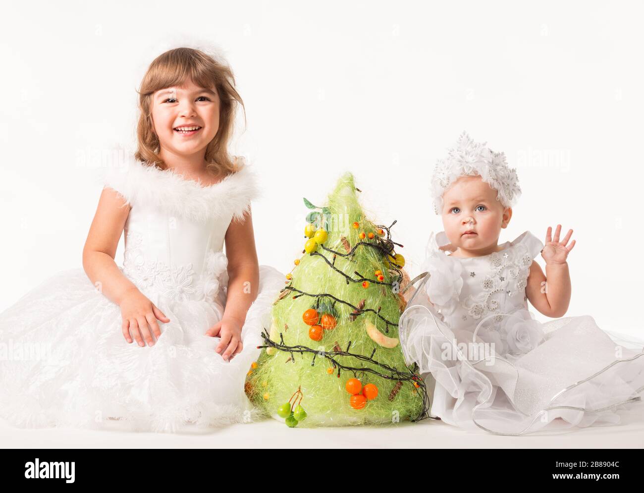 Vue sur le devant de jolis enfants mettant des guirlandes et des fruits sur  un arbre artificiel en pin. Les petites filles portent de belles robes de  carnaval et des bandeaux pour
