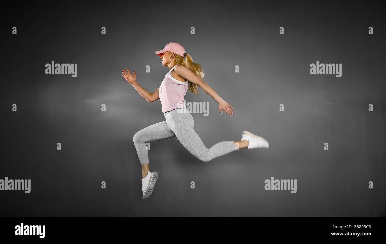 Jeune femme blonde avec corps ajusté sautant et courir sur fond gris. Modèle féminin dans l'exercice de vêtements de sport. Banque D'Images