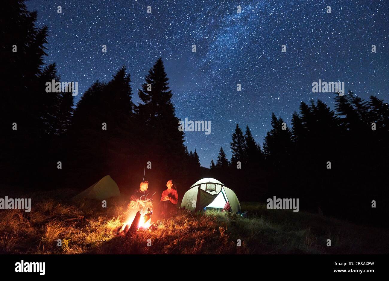 Vue sur l'avant de couples amoureux touristes assis par feu de camp lumineux près de deux tentes, profiter d'une belle nuit de camping ensemble sous le ciel sombre plein d'étoiles et la voie brillante lactée, chaud nuit d'été. Banque D'Images