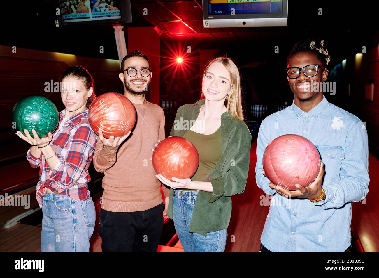 Quatre joueurs de bowling interculturel réussis avec des boules debout devant la caméra sur fond d'allées et d'écrans de score Banque D'Images