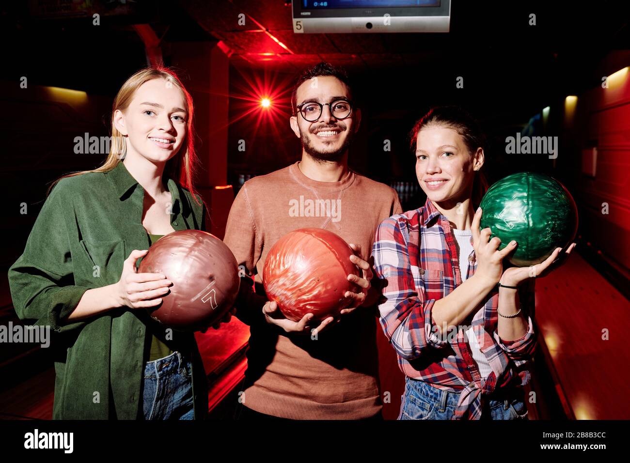 Trois jeunes joueurs de bowling interculturel et amicaux tenant des balles en se tenant dans la pièce sombre sur fond d'allées Banque D'Images