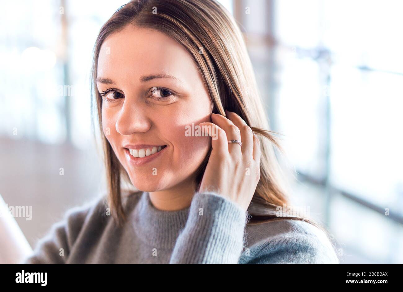 Belle jeune femme souriant et touchant les cheveux avec la main dans le bureau moderne ou le bâtiment d'affaires. Bonne personne regardant l'appareil photo. Banque D'Images