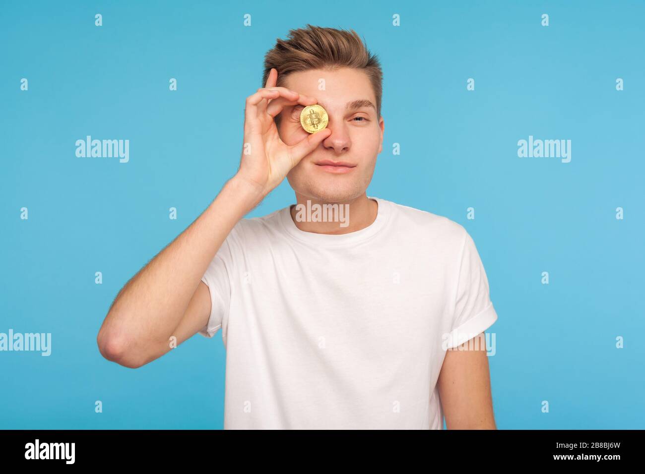 Portrait d'un homme riche et branché dans un t-shirt couvrant son œil avec un bitcoin doré, regardant un appareil photo avec une expression intelligente, confiant dans la crypto-monnaie Banque D'Images
