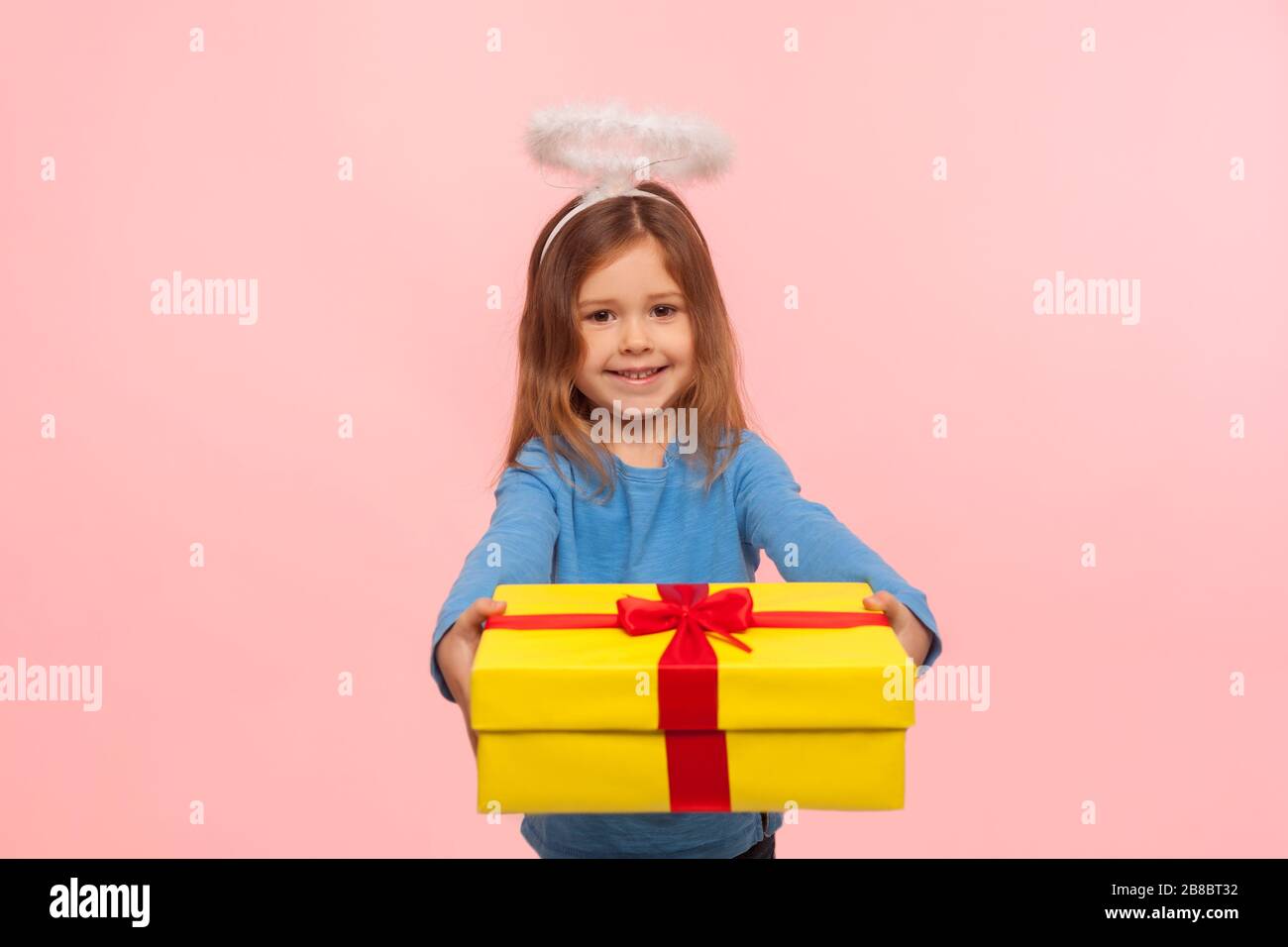 Portrait d'une généreuse petite fille angélique avec nimbus donnant un grand cadeau d'anniversaire à l'appareil photo et sourire, félicitant pour les vacances, le partage de cadeau. Banque D'Images