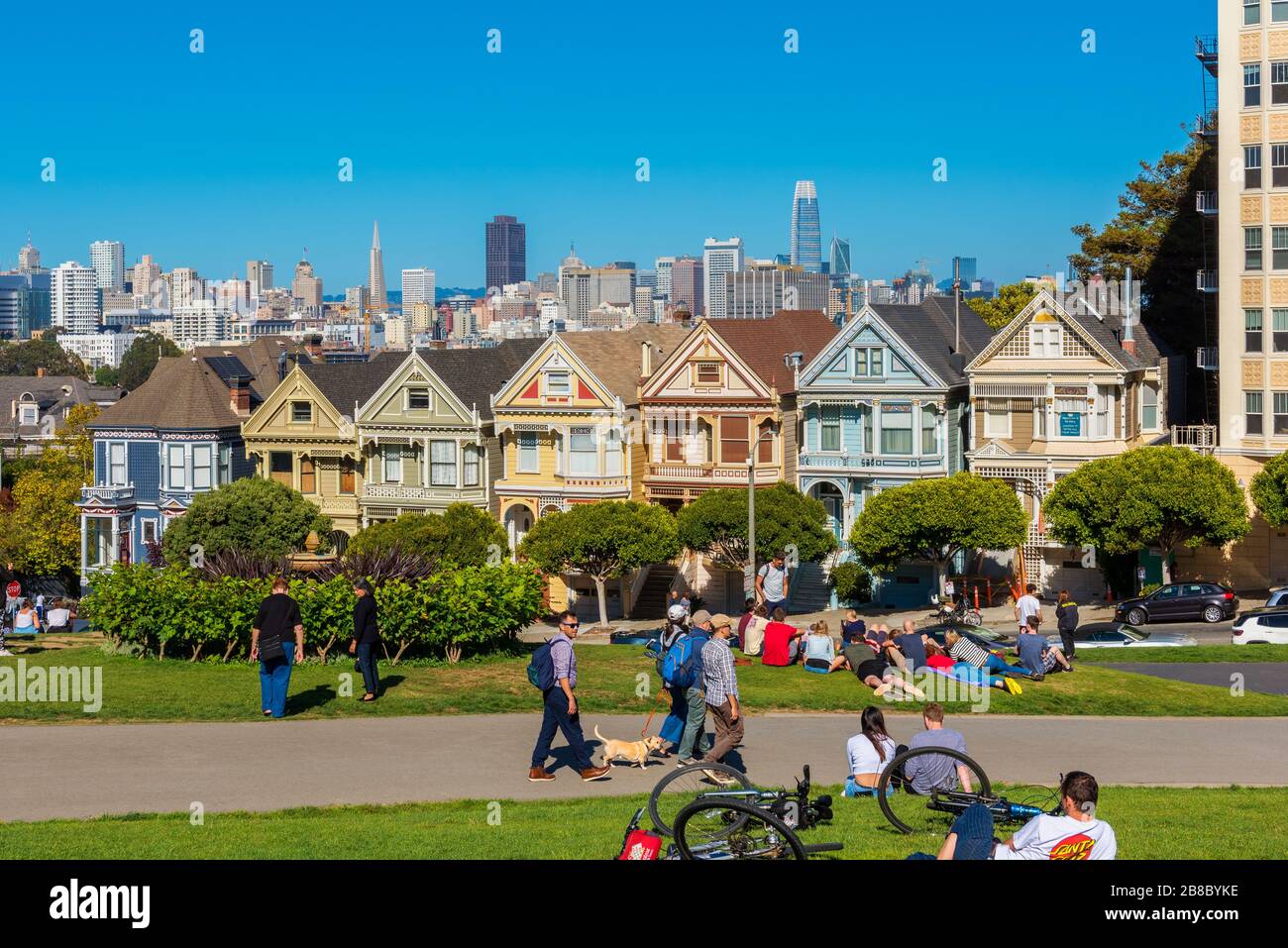 Les gens se détendant dans le parc devant les Maisons de femmes peintes à San Francisco, Californie, États-Unis. Les gratte-ciel de la ville sont visibles en arrière-plan. Banque D'Images