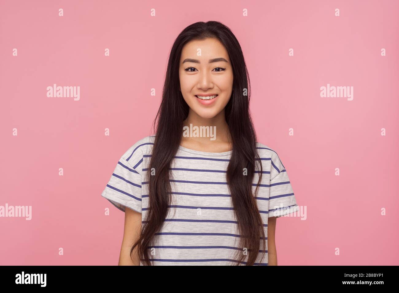 Portrait d'une jolie fille asiatique joyeuse avec de longs cheveux brunette dans un t-shirt rayé regardant un appareil photo avec un sourire toothy, en profitant de la vie heureuse, de bonne humeur. Banque D'Images