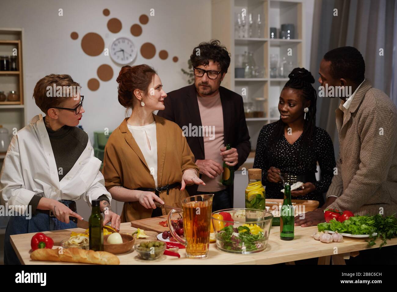 Groupe d'amis qui parlent et cuisent ensemble tout en se tenant dans la cuisine qu'ils préparent pour la fête Banque D'Images