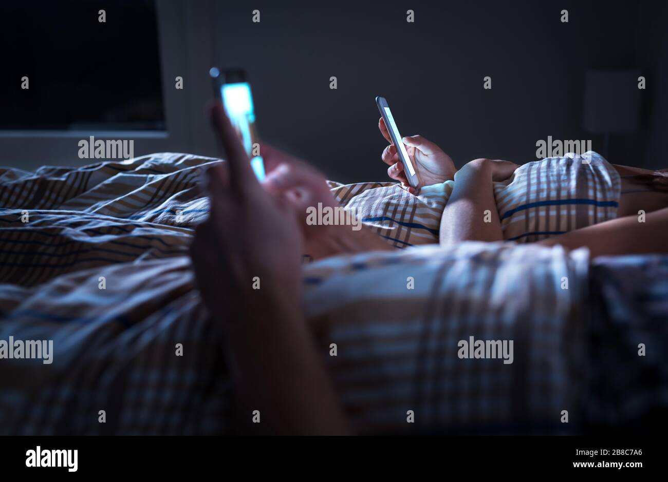 Un couple éloigné s'ennuyé s'ignorant l'un l'autre allongé dans le lit la nuit pendant que l'on utilise des téléphones mobiles. Accro au smartphone. Homme et femme obsédés et distraits. Banque D'Images