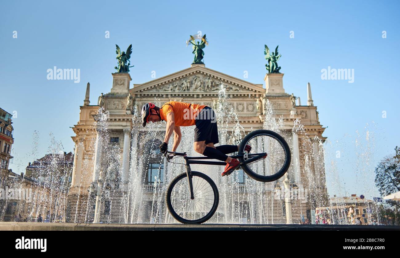 Vue avant du cycliste debout sur la roue avant du vélo tout terrain et regardant vers le bas. Le jeune athlète est en vélo au centre-ville près de la fontaine. Concept d'actif. Banque D'Images