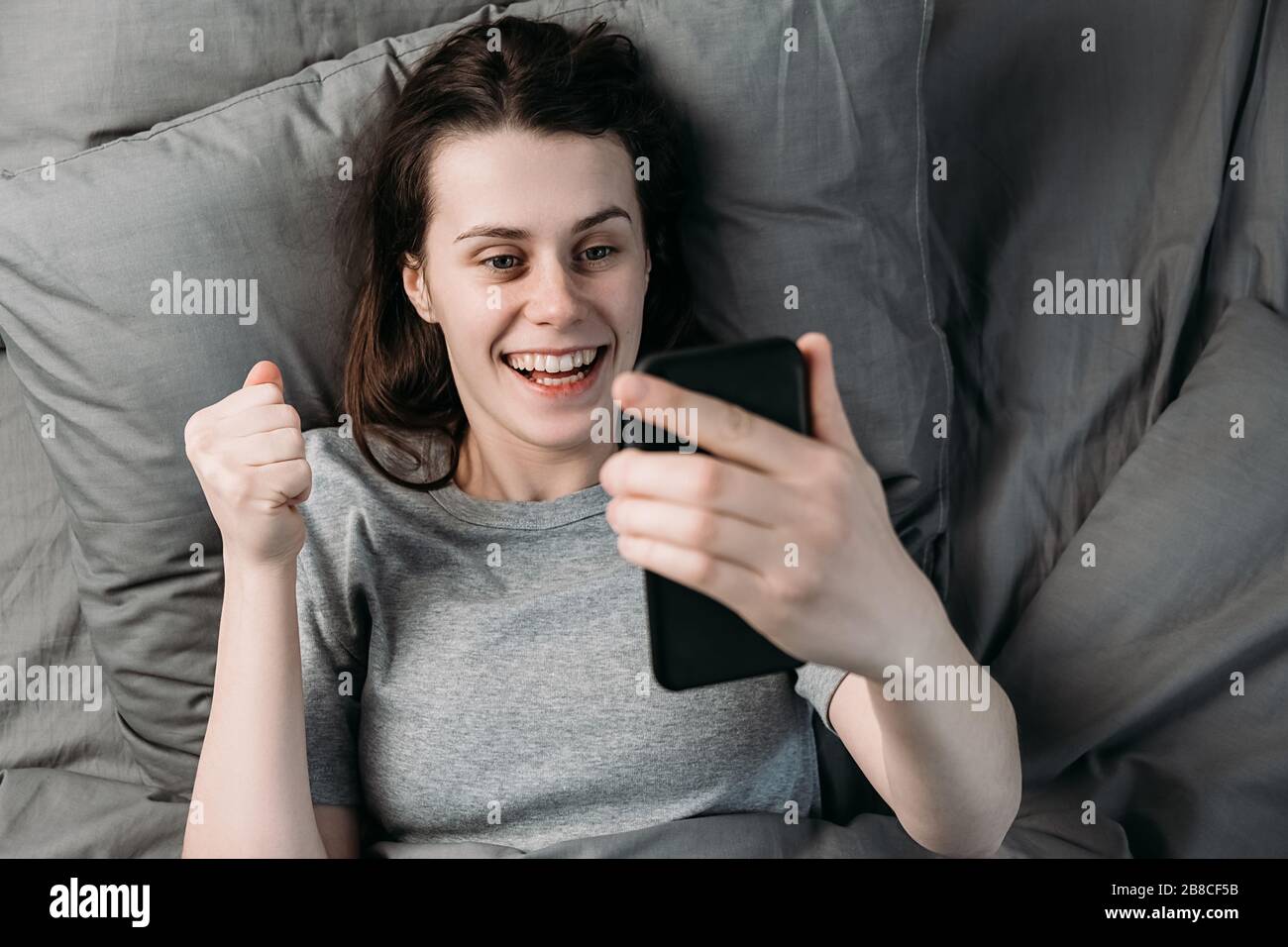 Vue de dessus de la jeune femme éveillée excitée se trouve dans le lit tenant dans le cri de smartphone main avec joie a reçu de grandes nouvelles le matin de début de journée. Concept Banque D'Images