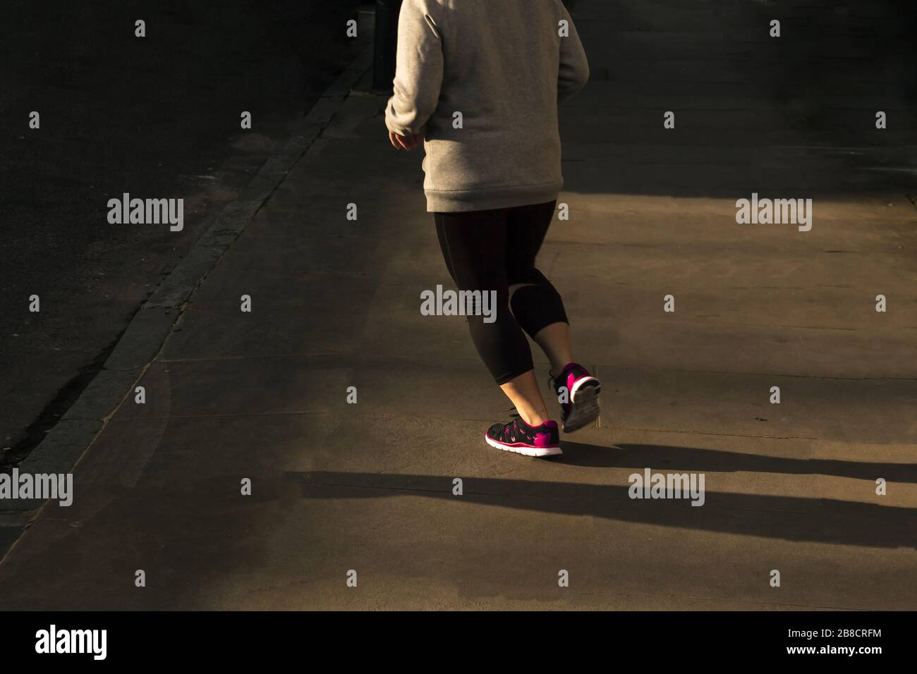 Vue arrière d'une femme en surpoids qui s'exécute dans un cadre lumineux sombre l'après-midi dans un cadre urbain et asphalté à la périphérie d'une rue de banlieue de la ville Banque D'Images