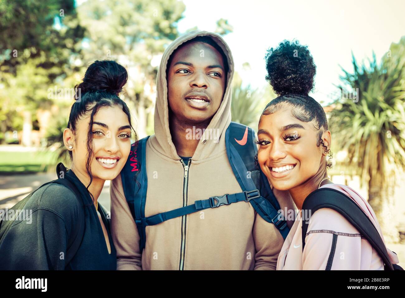 Un homme d'Afrique américain d'âge universitaire dans le hoodie entre deux coeds de couleur féminine avec des cool-dos à la mode sur le campus profitant de la vie d'étudiant Banque D'Images