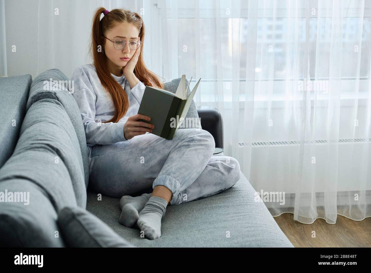 Une fille aux cheveux rouges avec des lunettes, avec de longues queues en pyjama assis sur le canapé et regarde bouleversé le livre, en pressant la paume au visage Banque D'Images