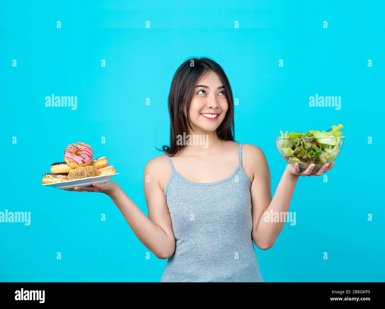 Jolie jeune femme asiatique tenant et choisissant entre un disque de beignets ou une salade de légumes dans un bol de verres sur fond bleu isolé, poids Banque D'Images