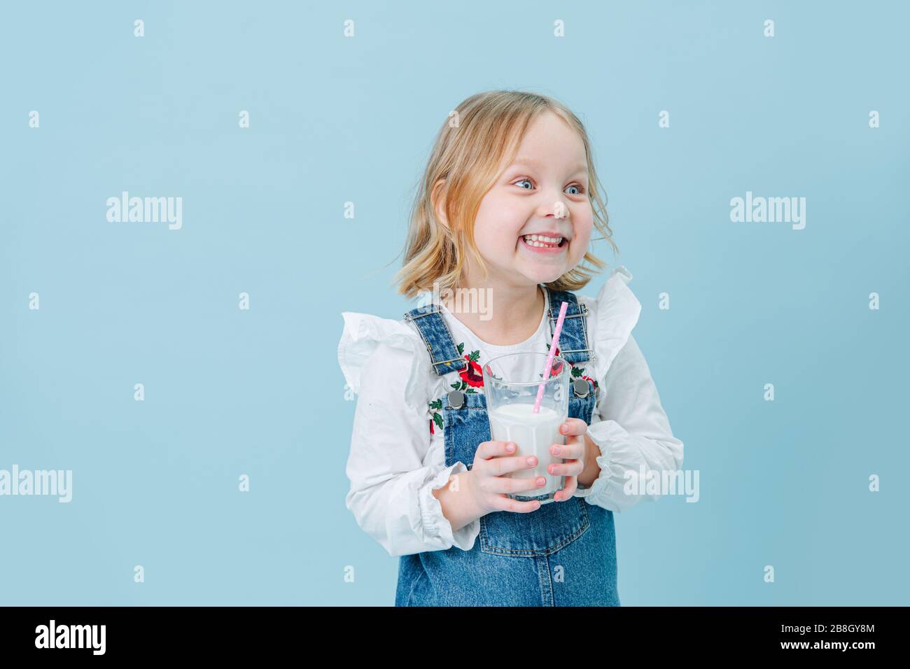 Drôle petite fille dans des salopettes avec un verre de milkshake sur fond bleu Banque D'Images