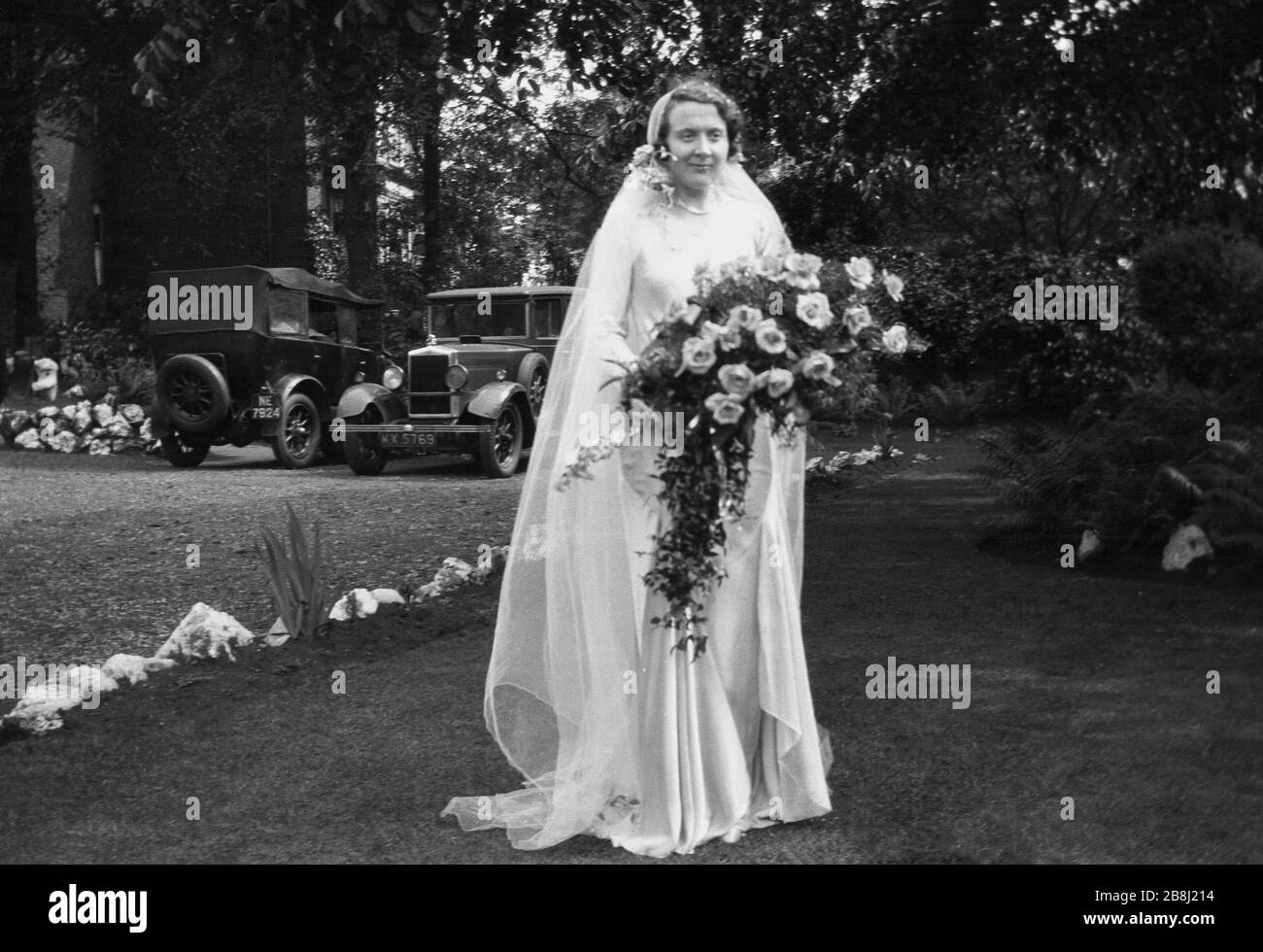 1920, historique, mariage, une mariée dans sa robe de mariage debout à  l'extérieur sur une pelouse posant pour une photo tenant un bouquet de  fleurs, avec des automobiles de l'époque garé sur