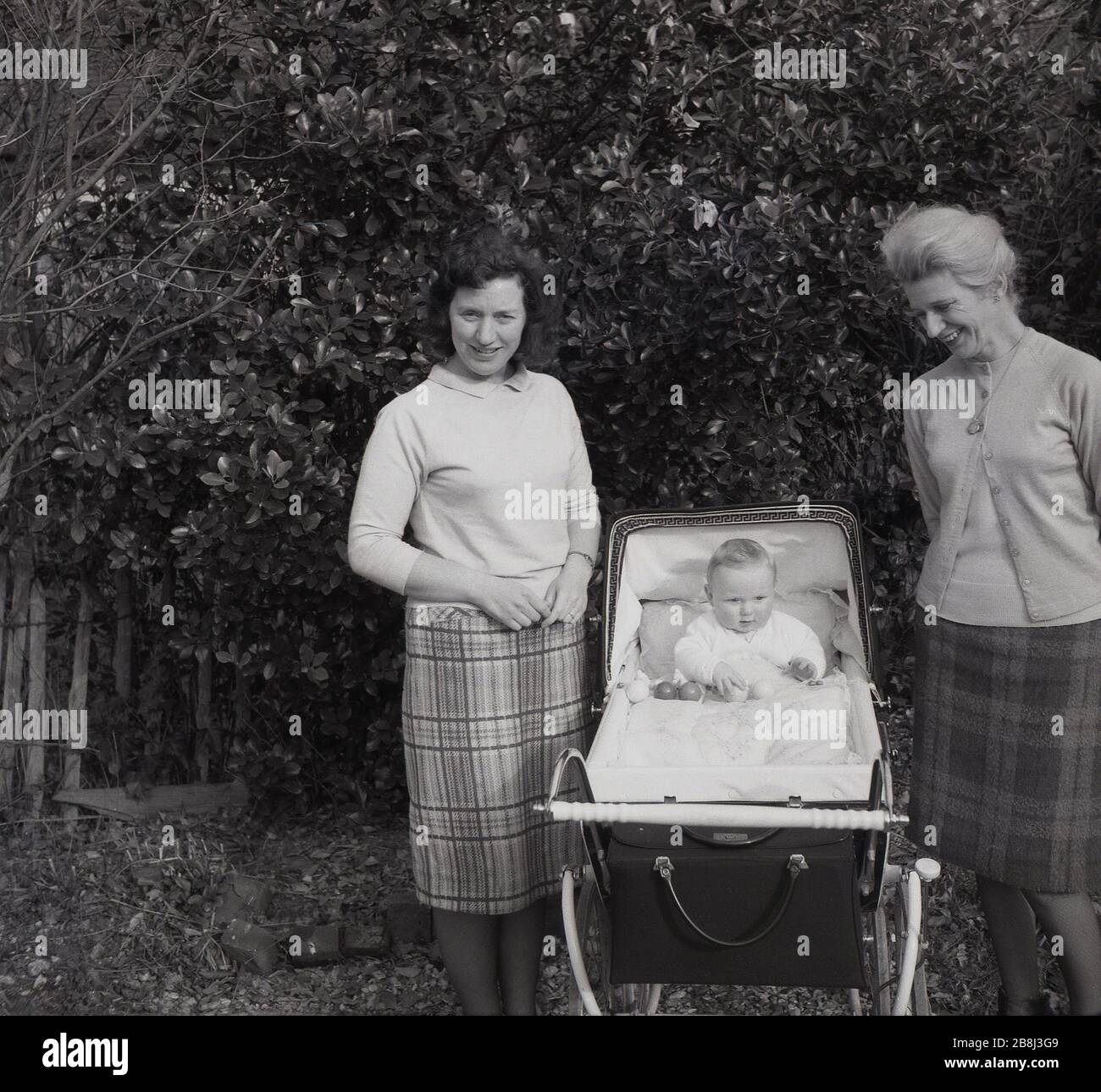 Années 50, historique, fière grand-mère, mère et bébé dans un pram de  l'époque debout ensemble à l'extérieur, Angleterre, Royaume-Uni. La mère et  le grand-mère de Bothe portent des jupes à carreaux ou
