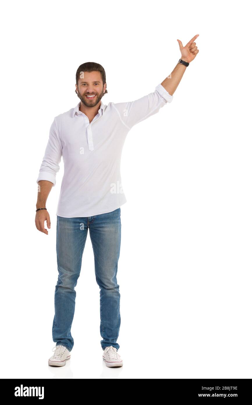 Beau jeune homme en Jean, baskets et chemise blanche est debout avec bras levé, regardant l'appareil photo, souriant et pointant vers le haut. Vue de face. Longueur totale Banque D'Images