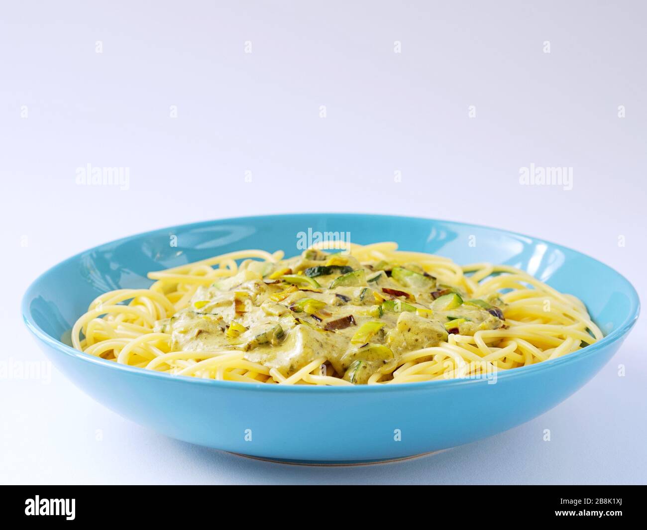 Spaghetti avec sauce crème avec courgettes et poireaux dans une assiette bleu clair sur fond blanc. Banque D'Images